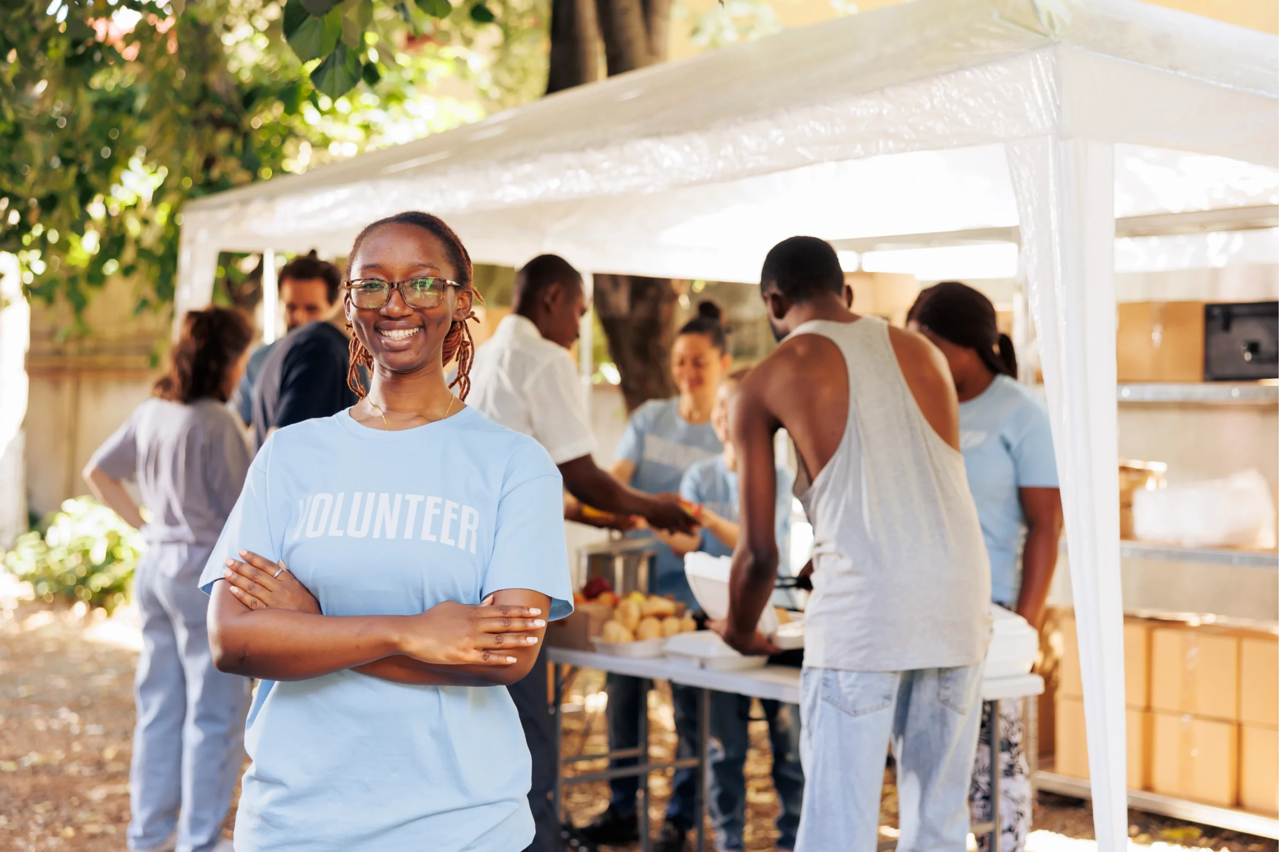 young_black_woman_with_glasses_stands_outdoors_arms_crossed_looking_camera_diverse_group_volunteers_supports_non_profit_program_dedicated_hunger_relief_helping_needy_individuals_1
